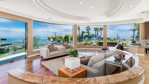 Ein großzügiger Wohnbereich mit einer gewölbten Marmorplatte, einer Couch und einem Couchtisch. Große Fenster bieten Blick auf das Meer und Palmen.
