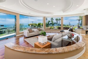 Ein großzügiger Wohnbereich mit einer gewölbten Marmorplatte, einer Couch und einem Couchtisch. Große Fenster bieten Blick auf das Meer und Palmen.