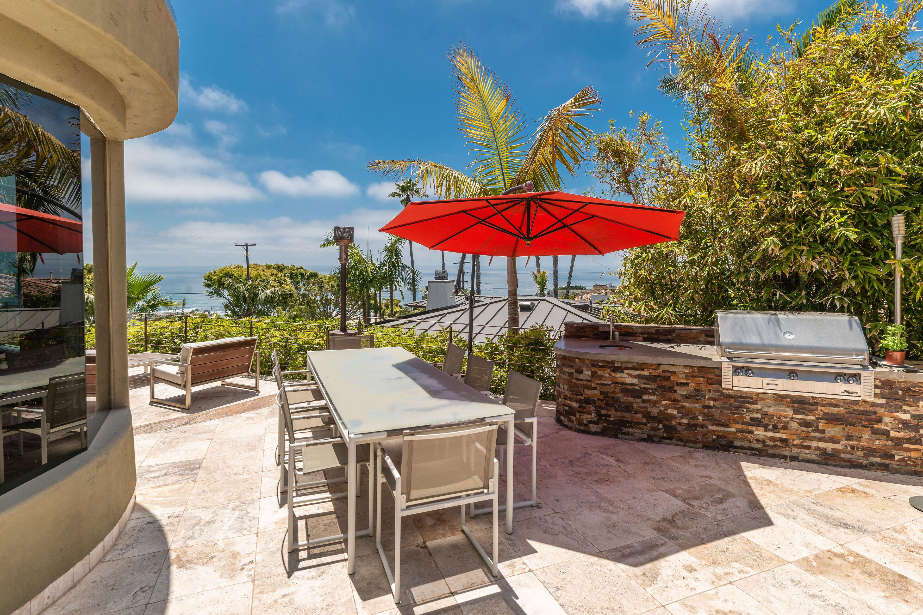 Um pátio externo com mesa e cadeiras, um guarda-sol vermelho e uma churrasqueira.