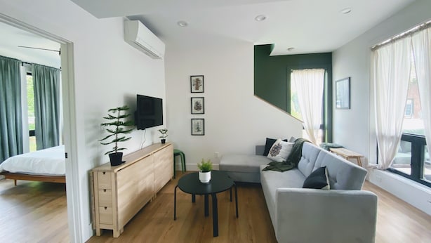 Una sala de estar moderna con un sofá, una mesita, un televisor montado en la pared y una cama al fondo.