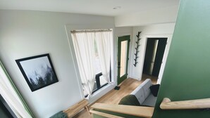Una escalera conduce a una habitación con paredes verdes, un banco de madera, un cuadro en la pared y una ventana con cortinas translúcidas.