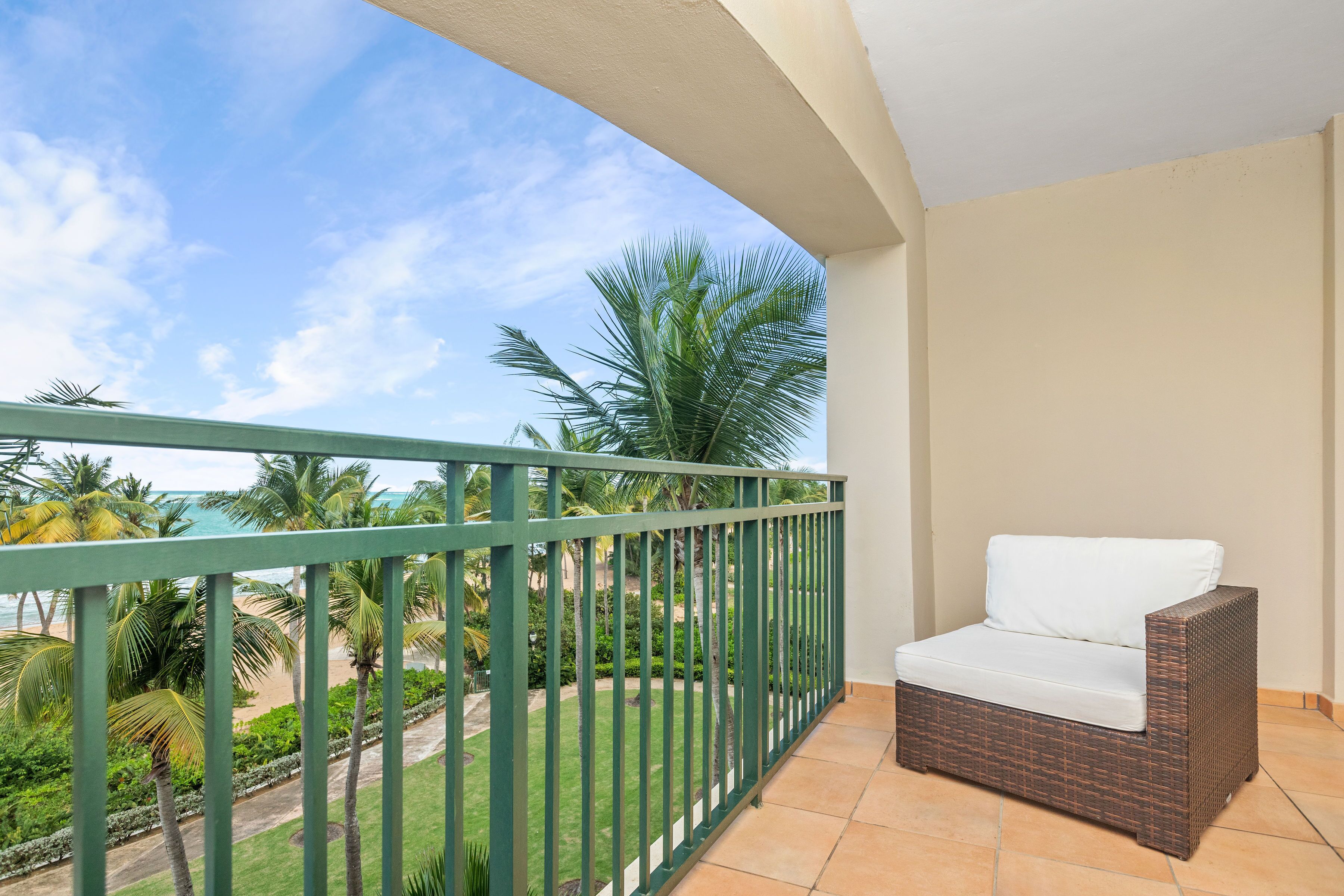 A balcony with a wicker sofa, a green metal railing, and a view of a beach and palm trees.