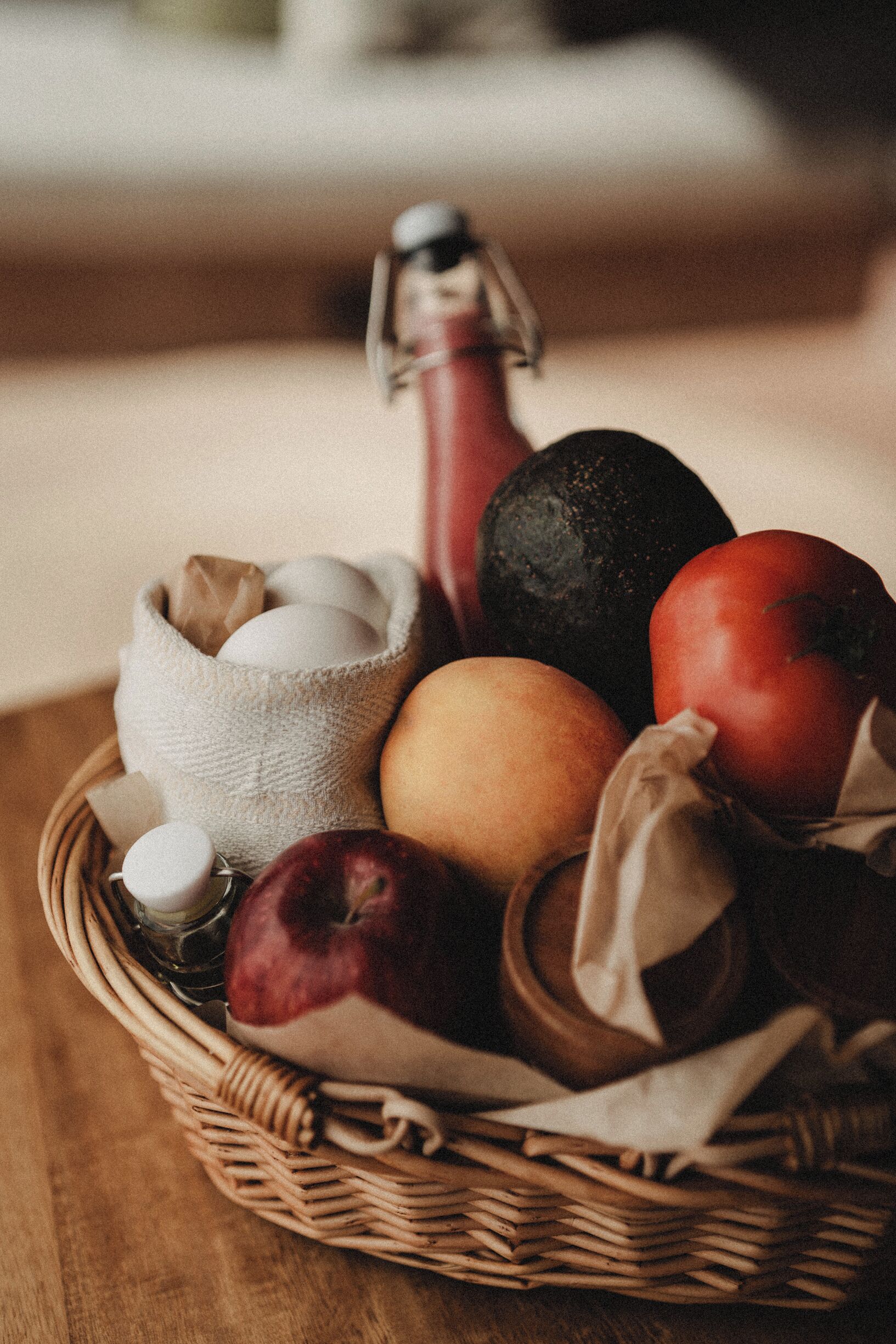 Una cesta de mimbre con huevos, una manzana roja, una manzana marrón y una fruta oscura, posiblemente aguacate, sobre una superficie de madera.