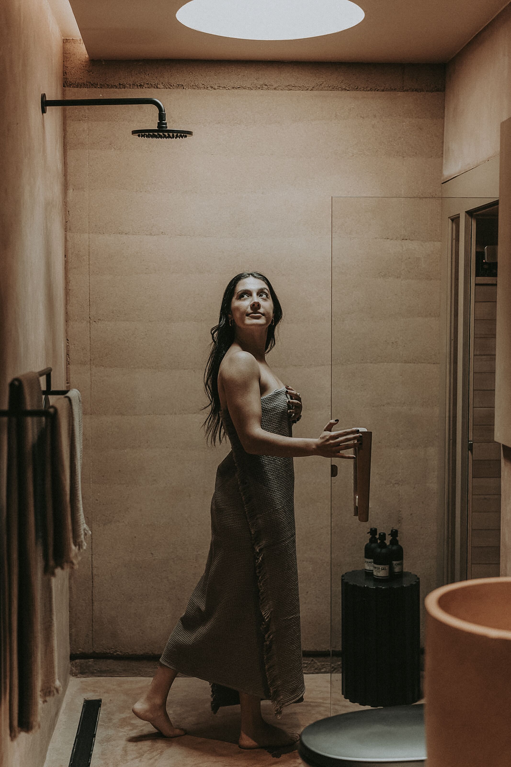 A woman in a bathroom with a towel rack, a showerhead, and a small table with bottles.