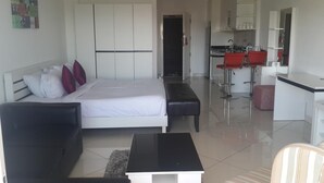 Una habitación de hotel moderna con una cama, un sofá negro, una mesita y una kitchenette con taburetes rojos.