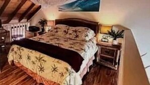 A bedroom with a wooden ceiling, a bed with a floral bedspread, a bedside table with a lamp, and a painting on the wall. - 1-bedroom condo in Maunaloa, just 10 minutes from the beach (Maunaloa)