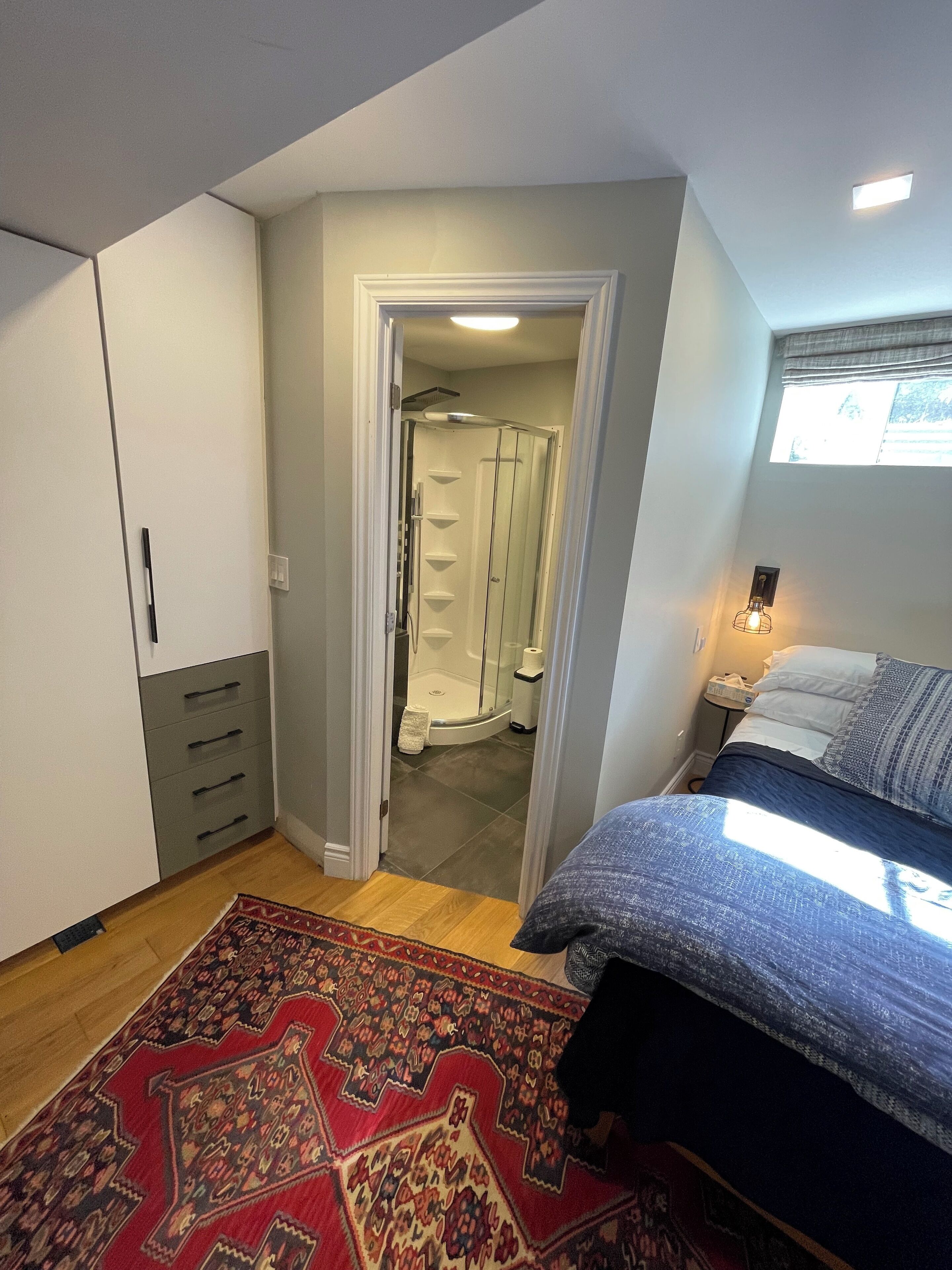A bedroom with a bed, a nightstand, a window, and a bathroom visible through an open door.