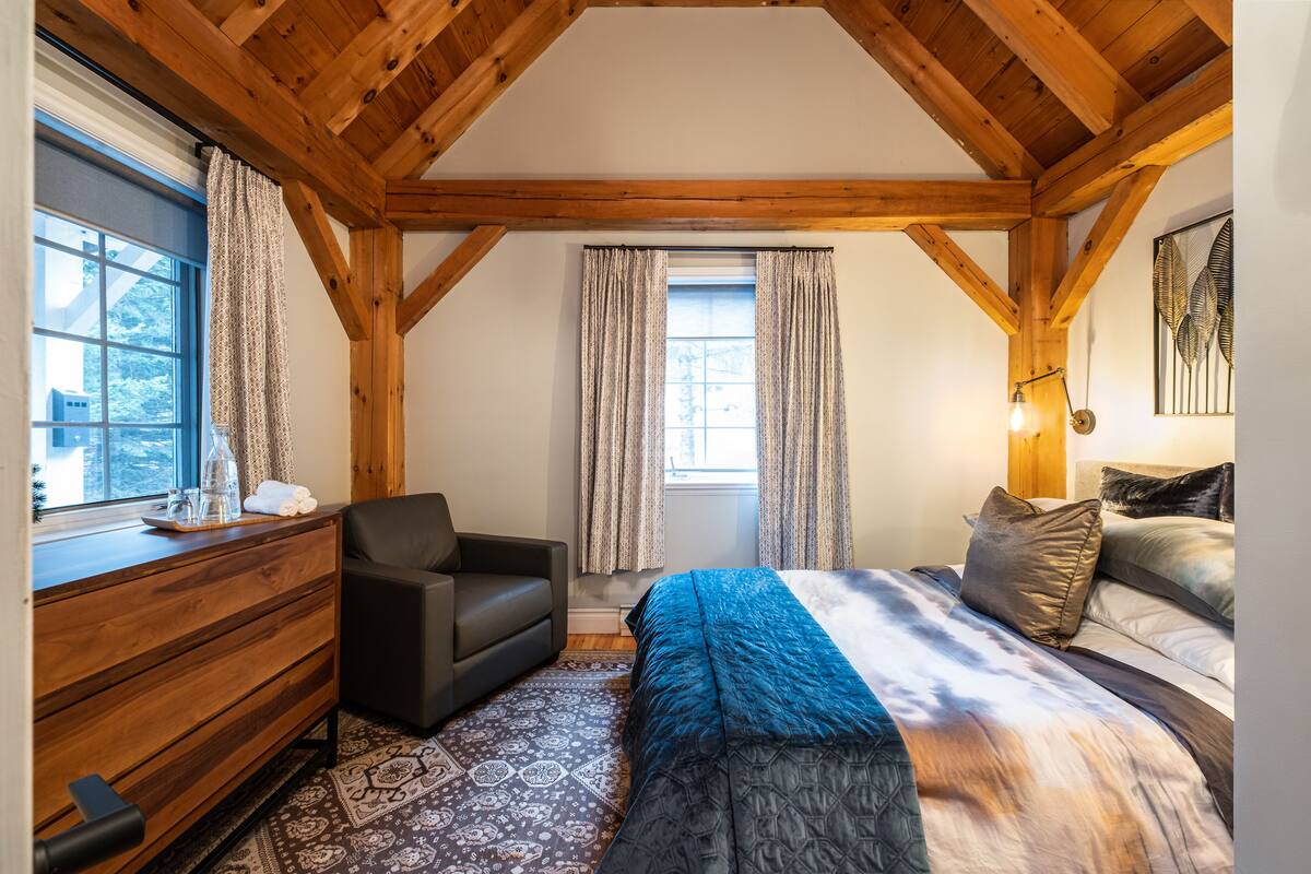 A bedroom with a wooden ceiling, a bed with a blue and white bedspread, a black sofa, a wooden dresser, and patterned rugs.
