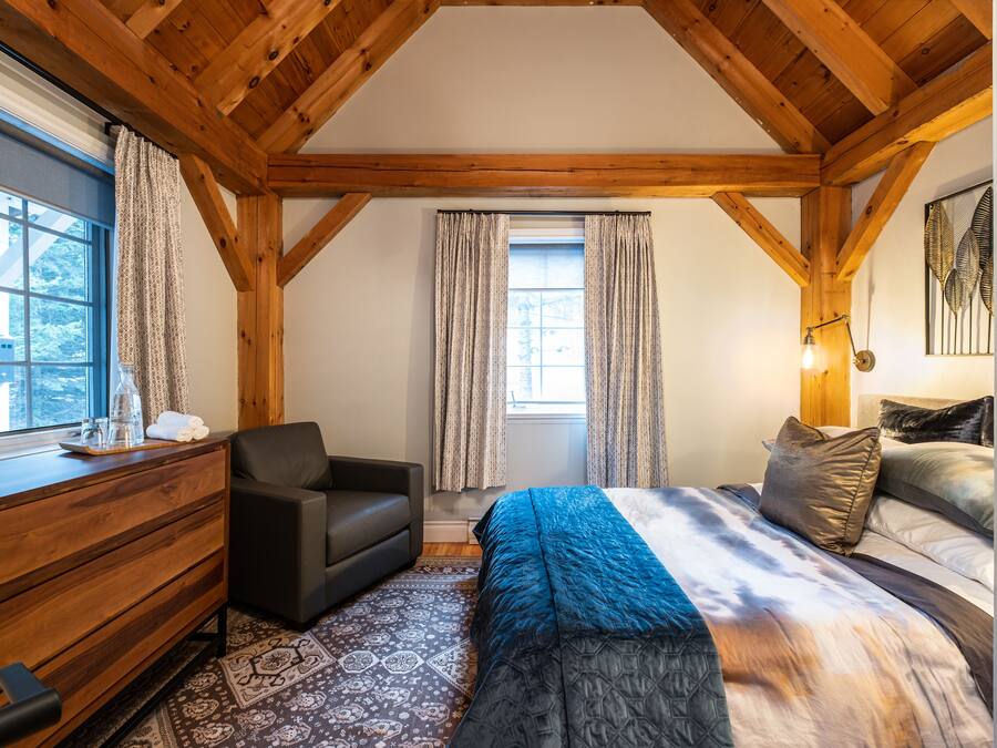 A bedroom with a wooden ceiling, a bed with a blue and white bedspread, a black sofa, a wooden dresser, and patterned rugs.