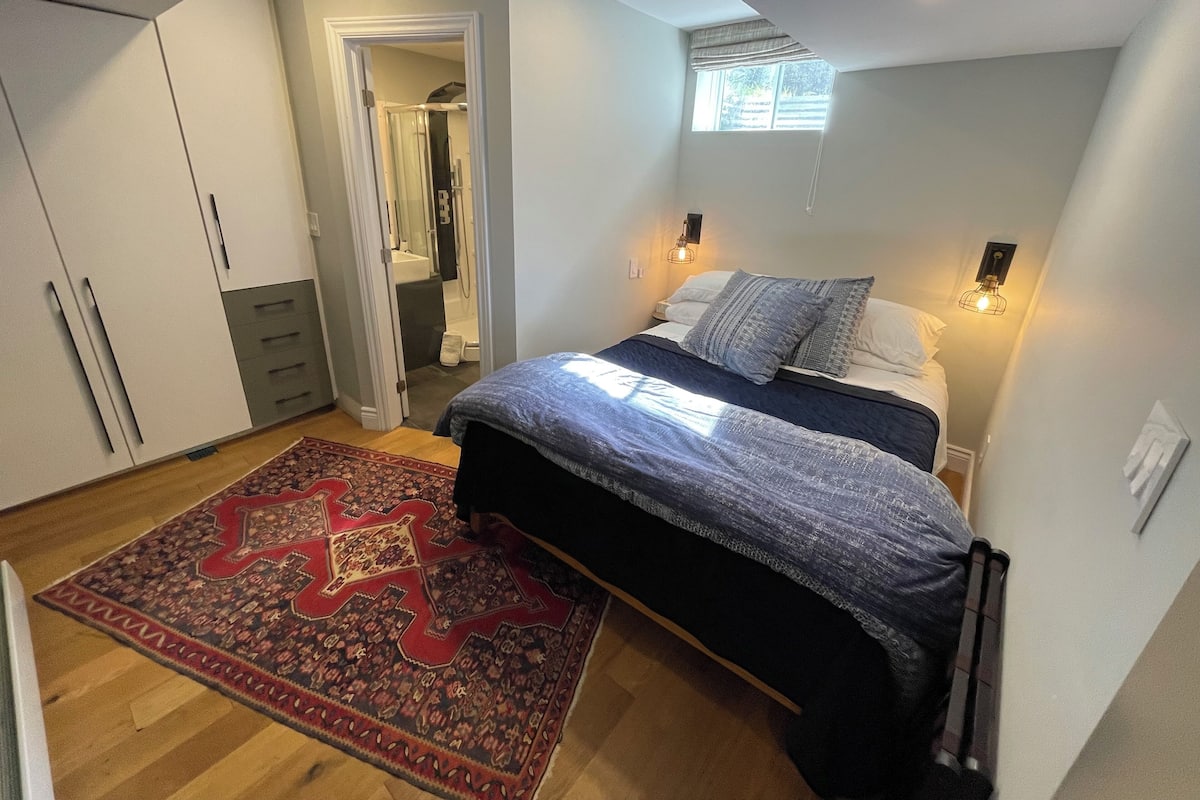 A bedroom with a bed, a red and black patterned rug, a wardrobe, and a bathroom visible through an open door.
