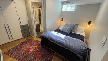 Un dormitorio con cama, alfombra estampada en rojo y negro, armario y baño visible a través de una puerta abierta.