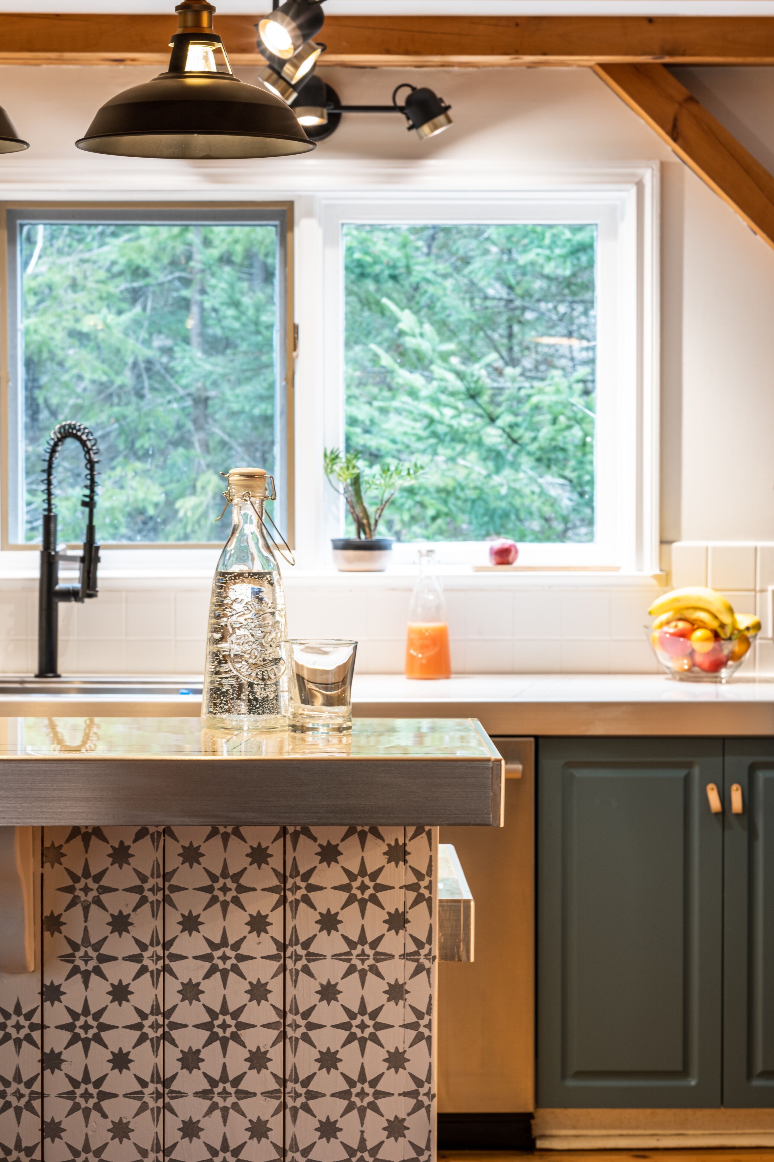 A kitchen with a window, a bottle, a glass, and a fruit bowl.