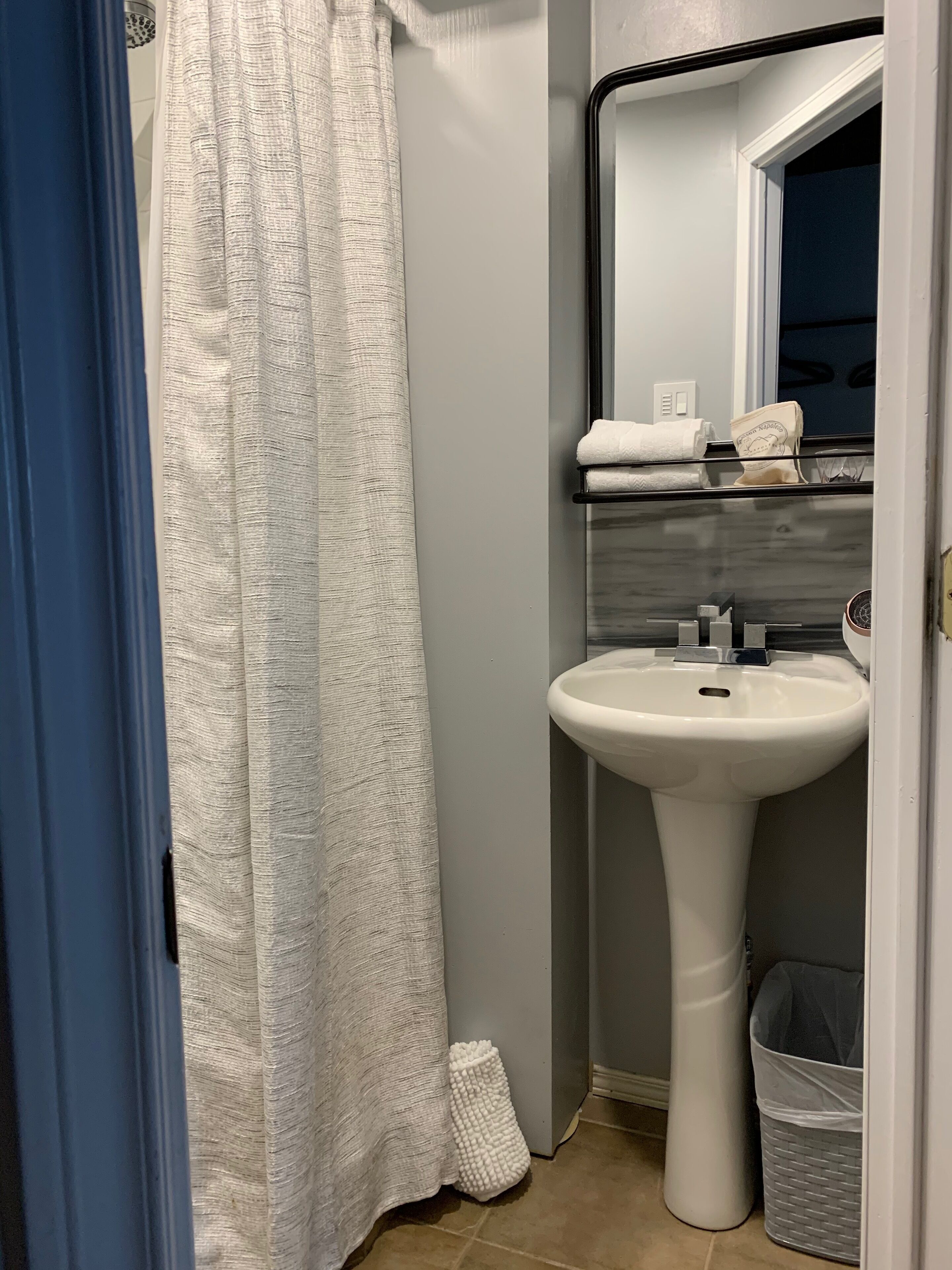 Un baño con cortina de ducha blanca, lavamanos blanco, espejo sobre el lavamanos y un cubo de basura.