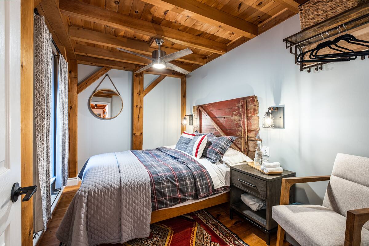 A bedroom with a wooden ceiling, a bed with a plaid bedspread, a nightstand, a chair, and a hanging light fixture.