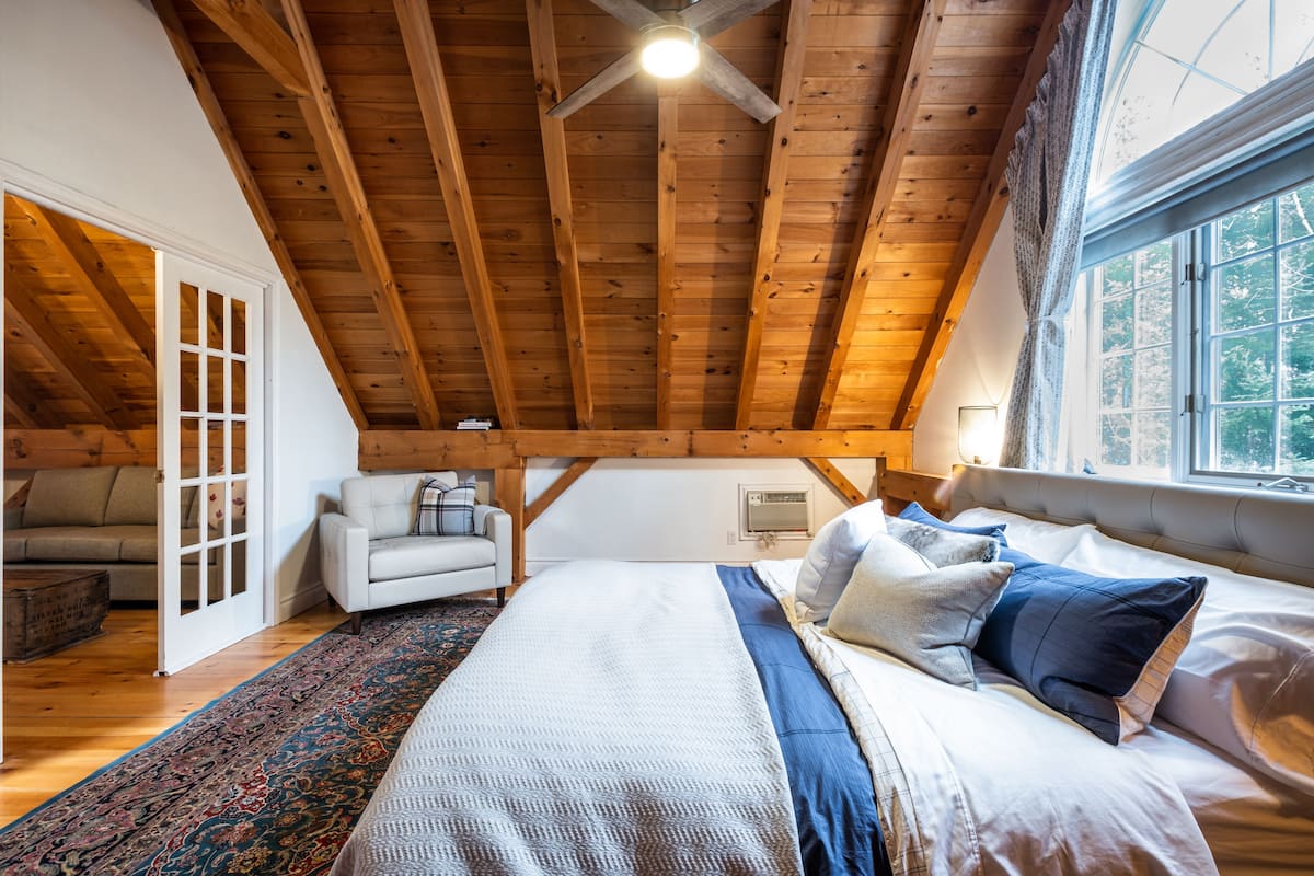 A bedroom with a wooden ceiling, a large bed with multiple pillows, a window with curtains, and a small table with a lamp.