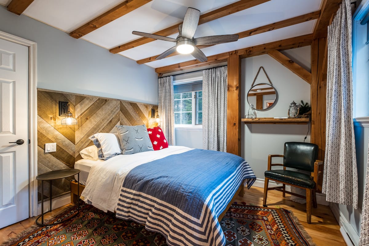 A bedroom with a bed, a chair, a desk, a ceiling fan, a window with curtains, and a wooden wall panel.