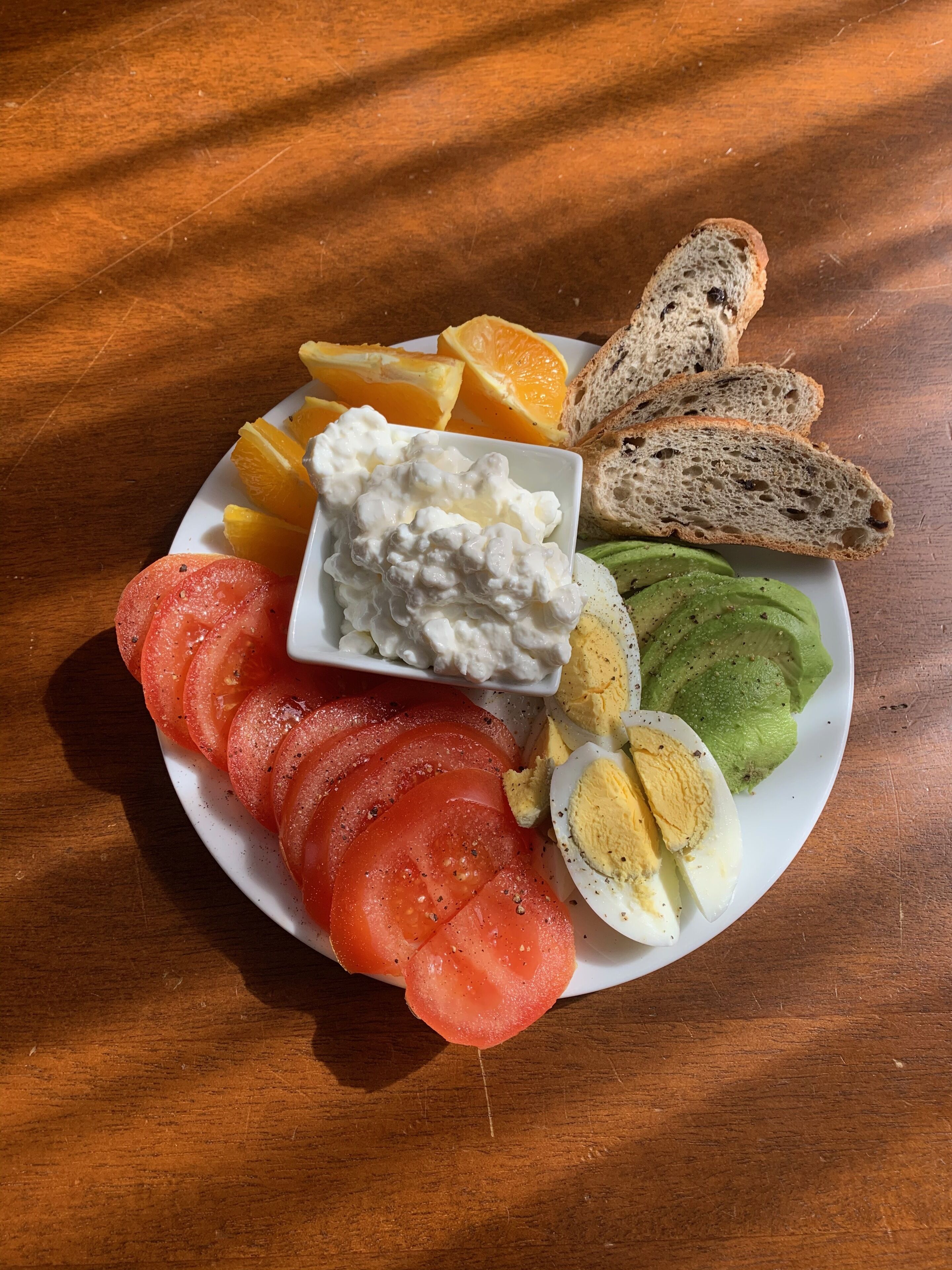 A plate with sliced tomatoes, eggs, avocado, orange slices, and a small bowl of yogurt.