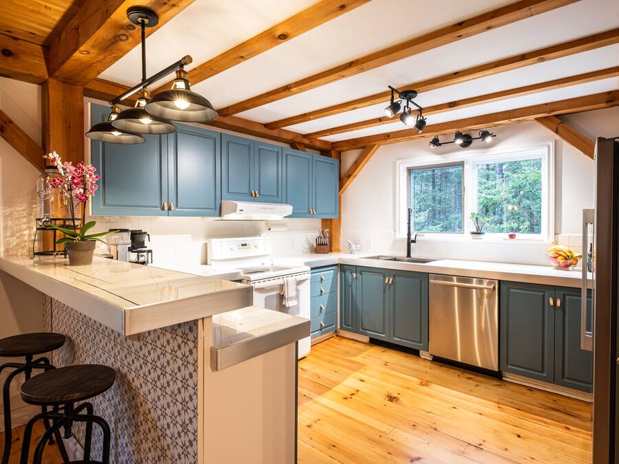 Una cocina con vigas de madera, armarios azules, encimera blanca y una ventana con vistas a árboles.