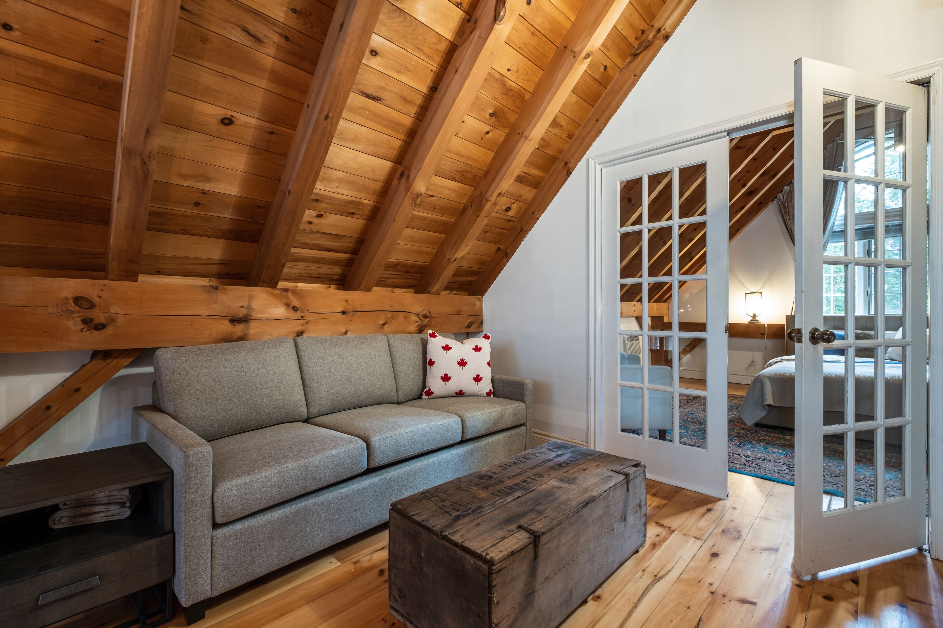 A cozy room with a wooden ceiling, a gray sofa with a red and white pillow, and a wooden chest of drawers.