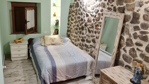 Un dormitorio acogedor con techo de madera, pared de piedra, cama con manta a rayas, cómoda y una mesita.