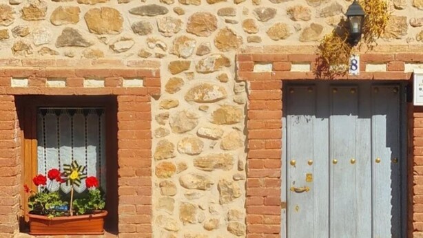 Un edificio de piedra con aspecto rústico, que presenta un letrero azul con letras blancas, una puerta de madera con tiradores dorados y macetas con flores en los alféizares de las ventanas.