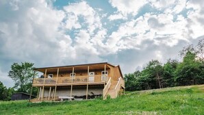 A two-story wooden house with a balcony on the upper floor, surrounded by greenery and under a partly cloudy sky.