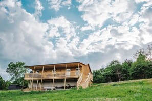 Une maison en bois à deux étages, dotée d’un balcon à l’étage supérieur, entourée de verdure et sous un ciel partiellement nuageux.