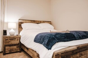Une chambre à coucher avec un cadre de lit en bois, du linge de lit blanc, une couverture bleu foncé, une table de chevet avec une lampe et une commode.
