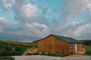 Une cabane en bois dotée d’un porche et d’un escalier, entourée d’une zone recouverte de gravier et d’une végétation luxuriante, sous un ciel nuageux.