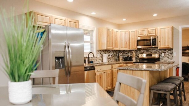A modern kitchen with wooden cabinets, stainless steel appliances, a granite countertop, and a dining area with a white table and two chairs.