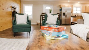 A cozy living room with a green armchair, a wooden coffee table with board games, and a kitchen in the background.