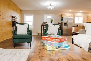 Un salon chaleureux avec un fauteuil vert, une table basse en bois sur laquelle sont posés des jeux de société, et une cuisine en arrière-plan.