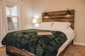 Quarto com cabeceira de madeira, colcha verde com textura, travesseiros brancos e abajur.