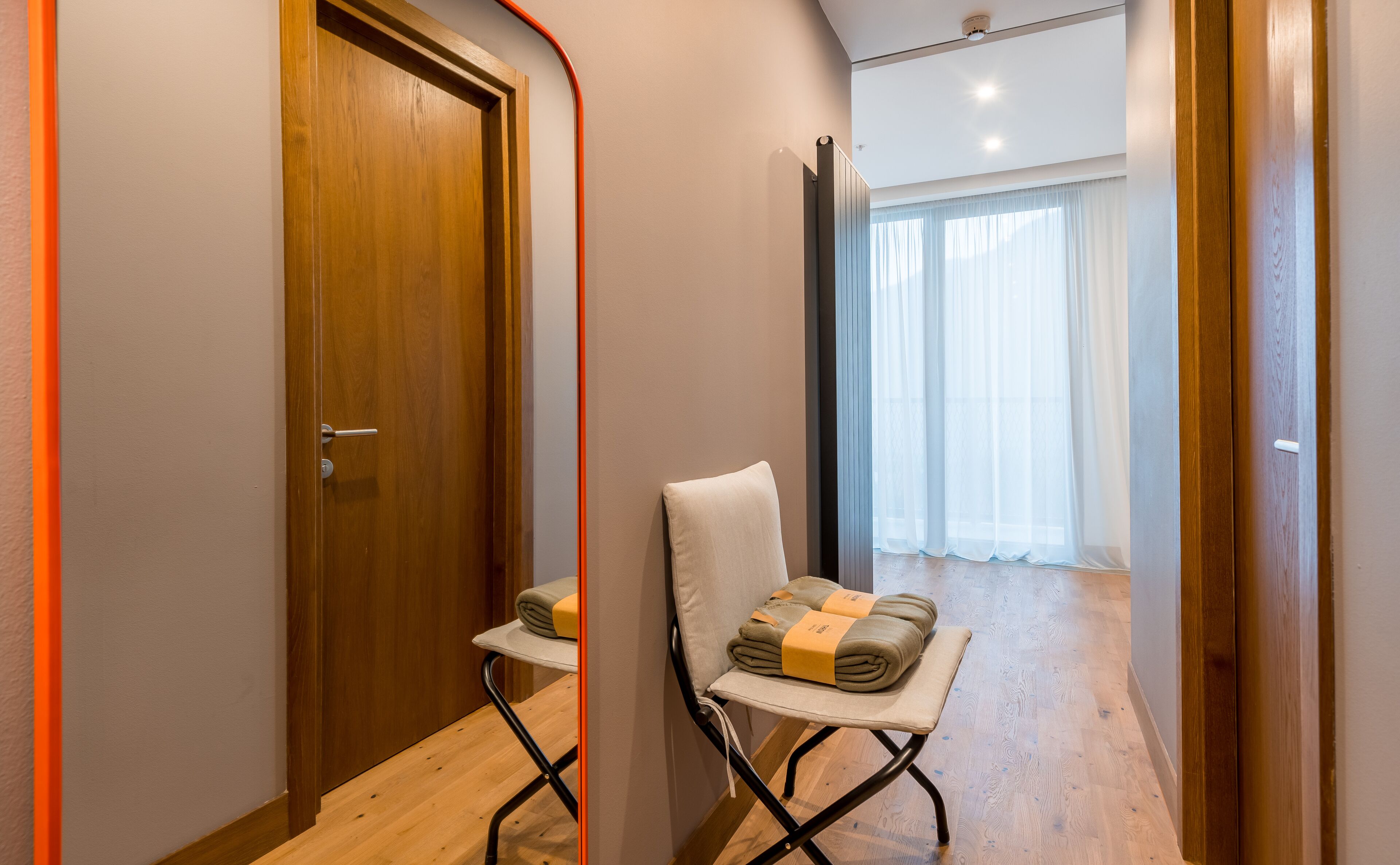 A narrow hallway with wooden doors, a folding chair with towels, and a view of a room with a large window and curtains.