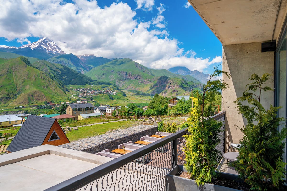 Un balcon avec un mur en pierre, des sièges d’extérieur et une vue sur une chaîne de montagnes et un village.