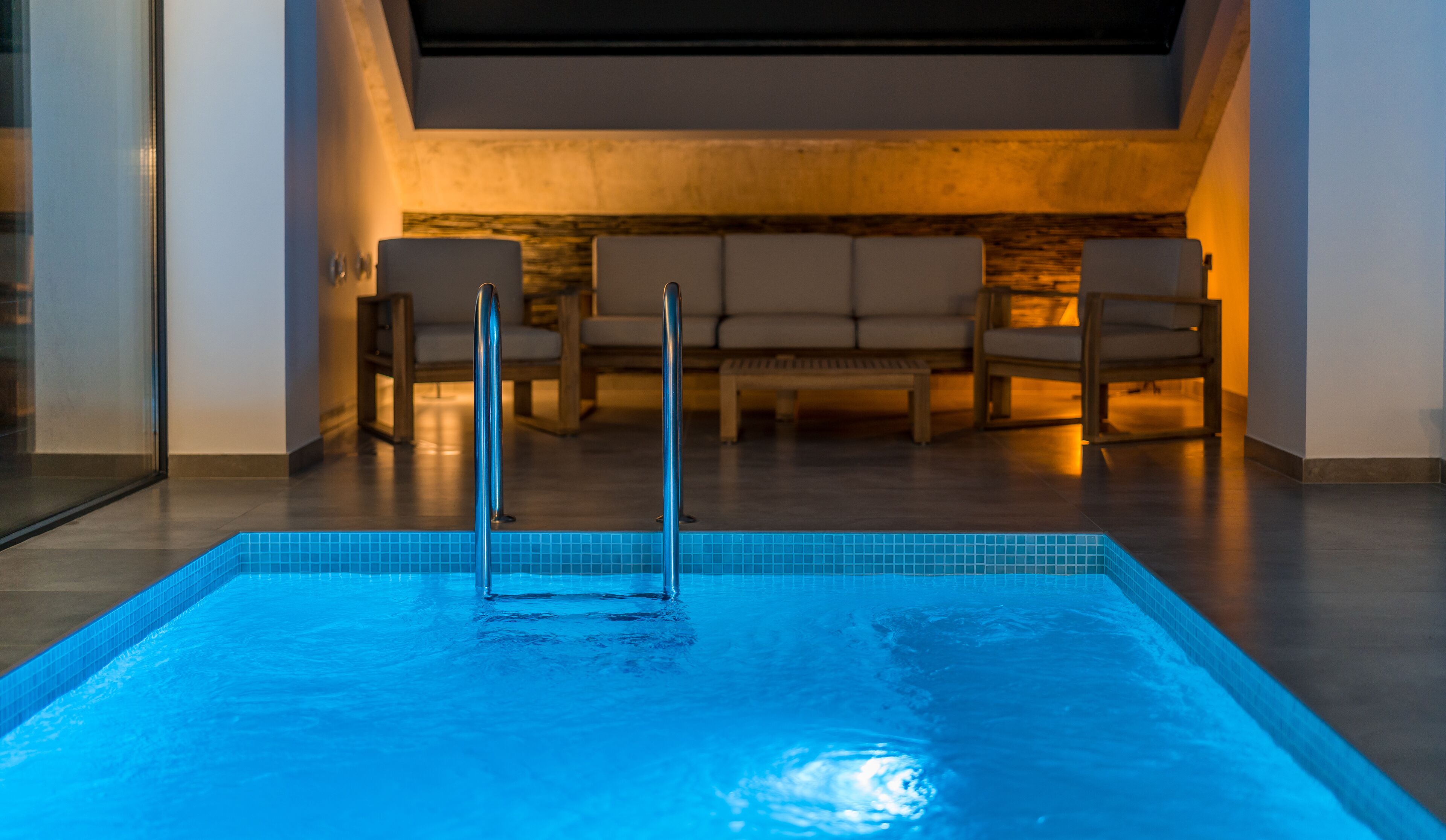 A swimming pool with a metal ladder, surrounded by a lounge area with chairs and a stone wall.