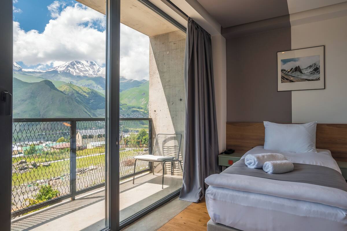 Une chambre d’hôtel avec un grand balcon, un lit avec du linge de maison blanc, un tableau au mur et une vue sur les montagnes.