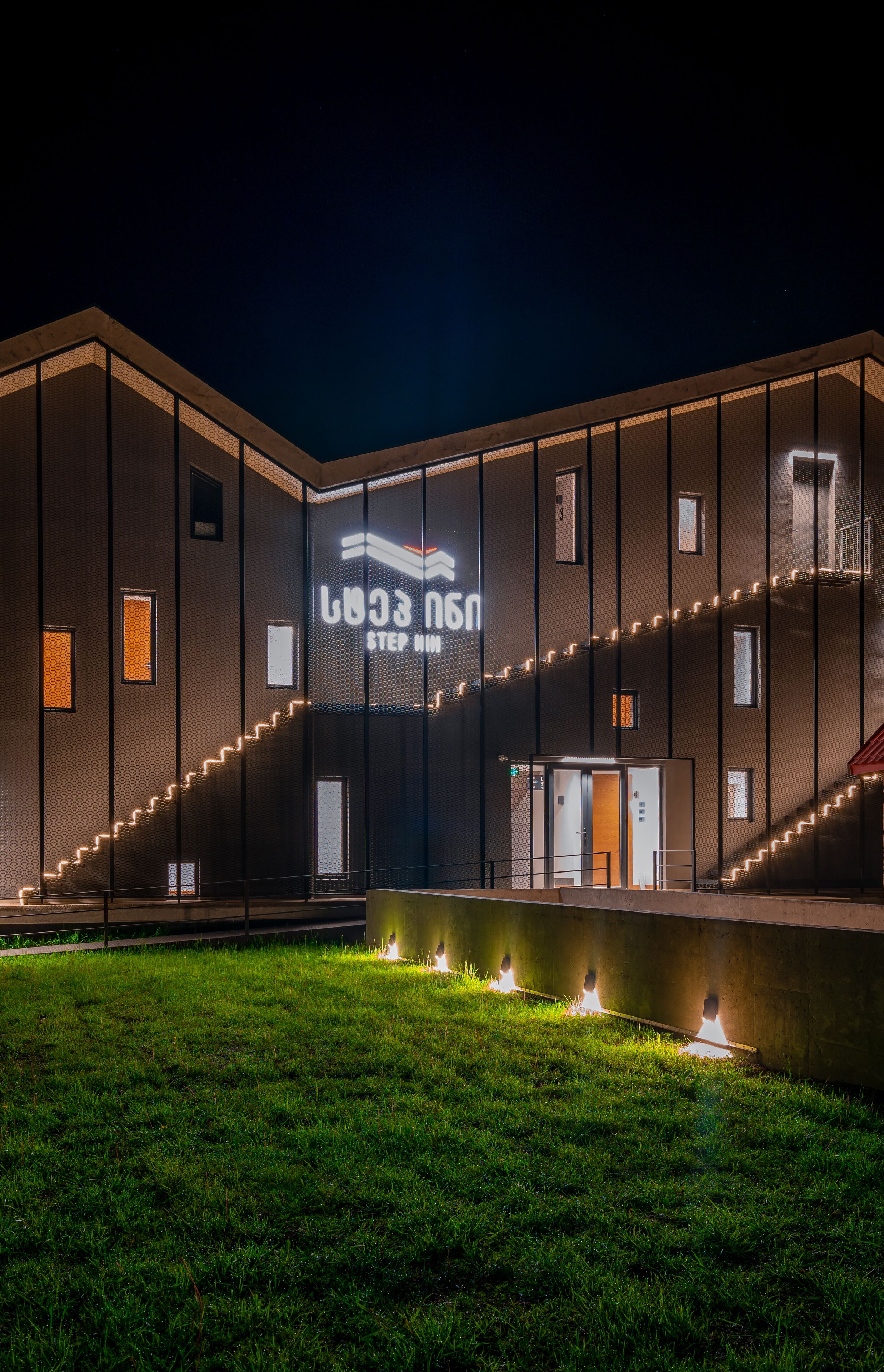Un edificio moderno con un gran letrero iluminado que dice "STEP MIM" y una escalera bien iluminada que conduce a la entrada.