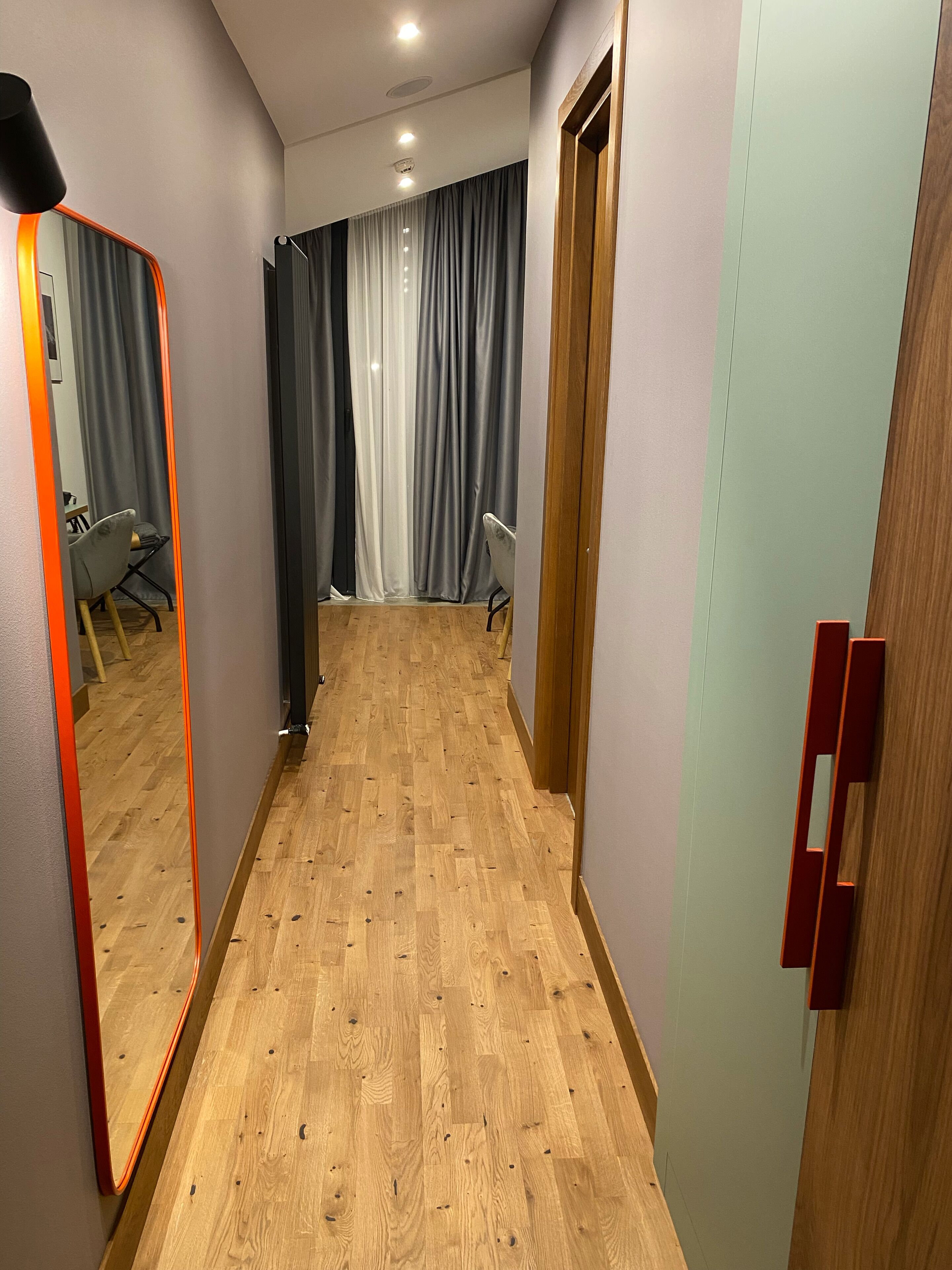 A hallway with wooden flooring, a mirror, and a door with a red handle.