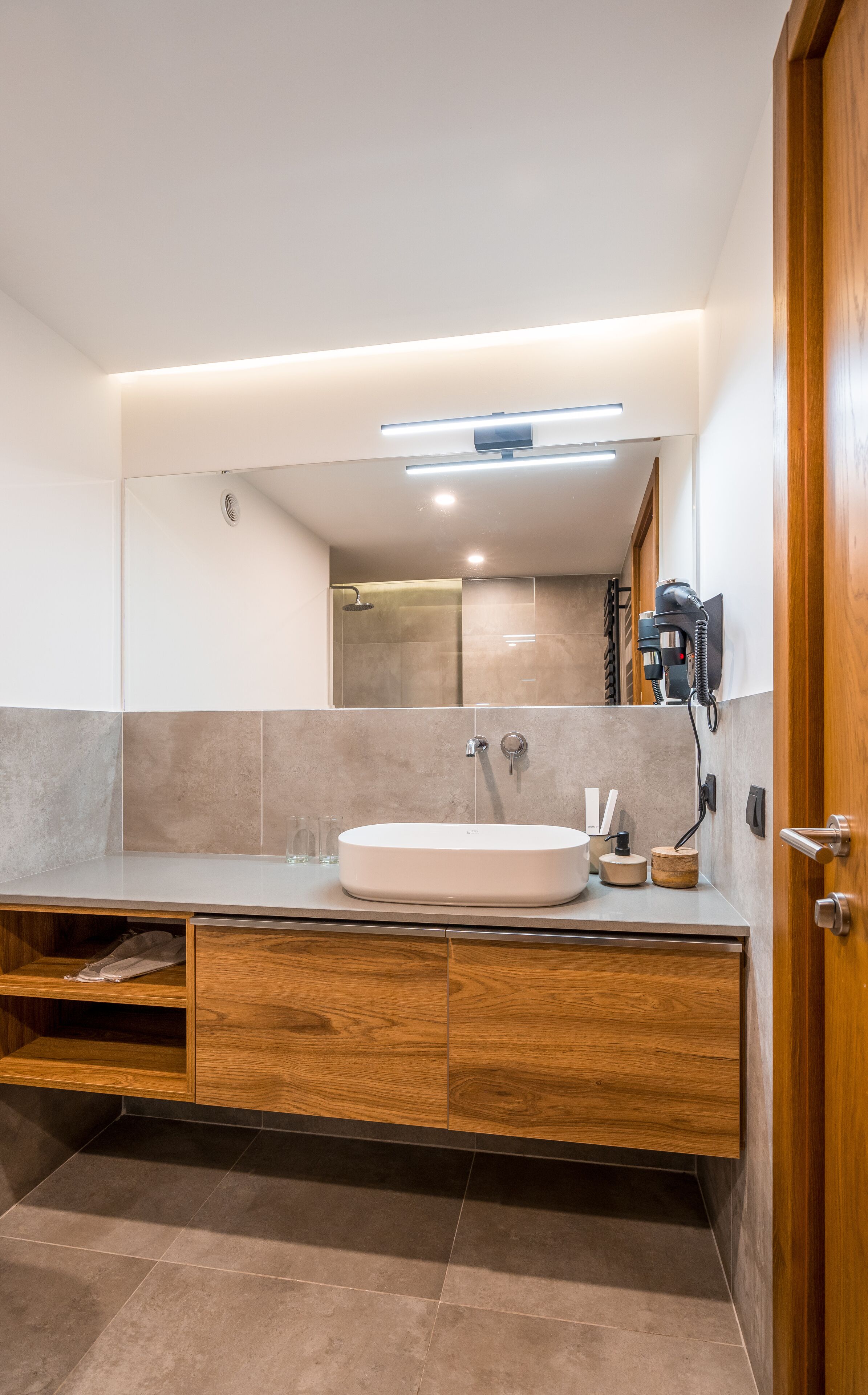 A modern bathroom with a large mirror, a round sink, wooden cabinets, and a hairdryer mounted on the wall.