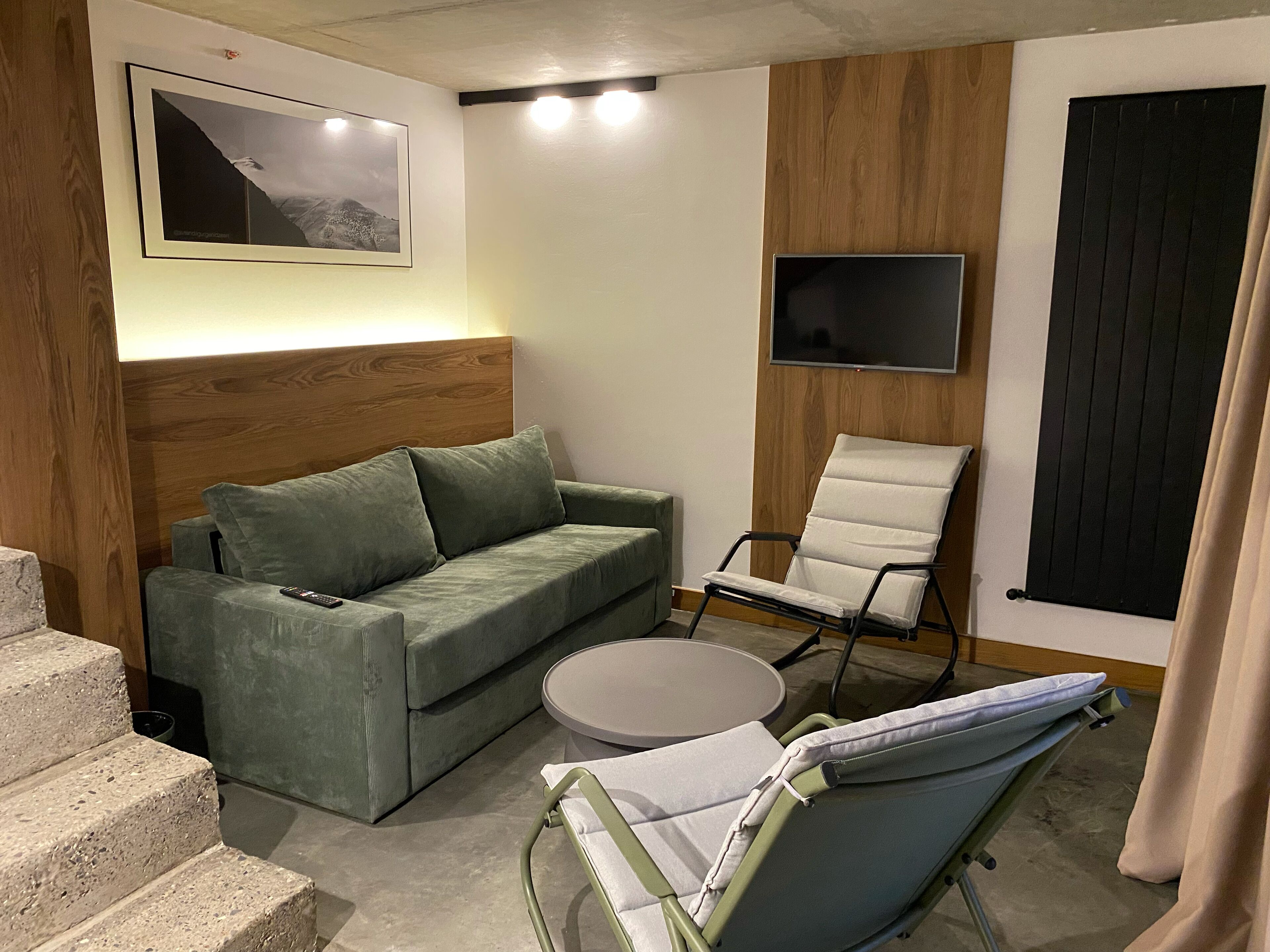 Un salon moderne avec un canapé vert, un fauteuil blanc, une petite table ronde et un téléviseur fix é au mur.