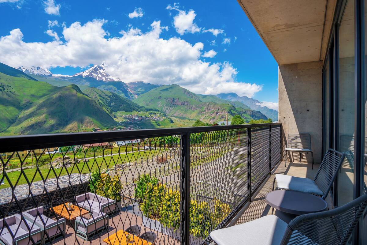 Un balcon avec des sièges d’extérieur, une vue sur des collines verdoyantes et des montagnes, et un ciel dégagé.