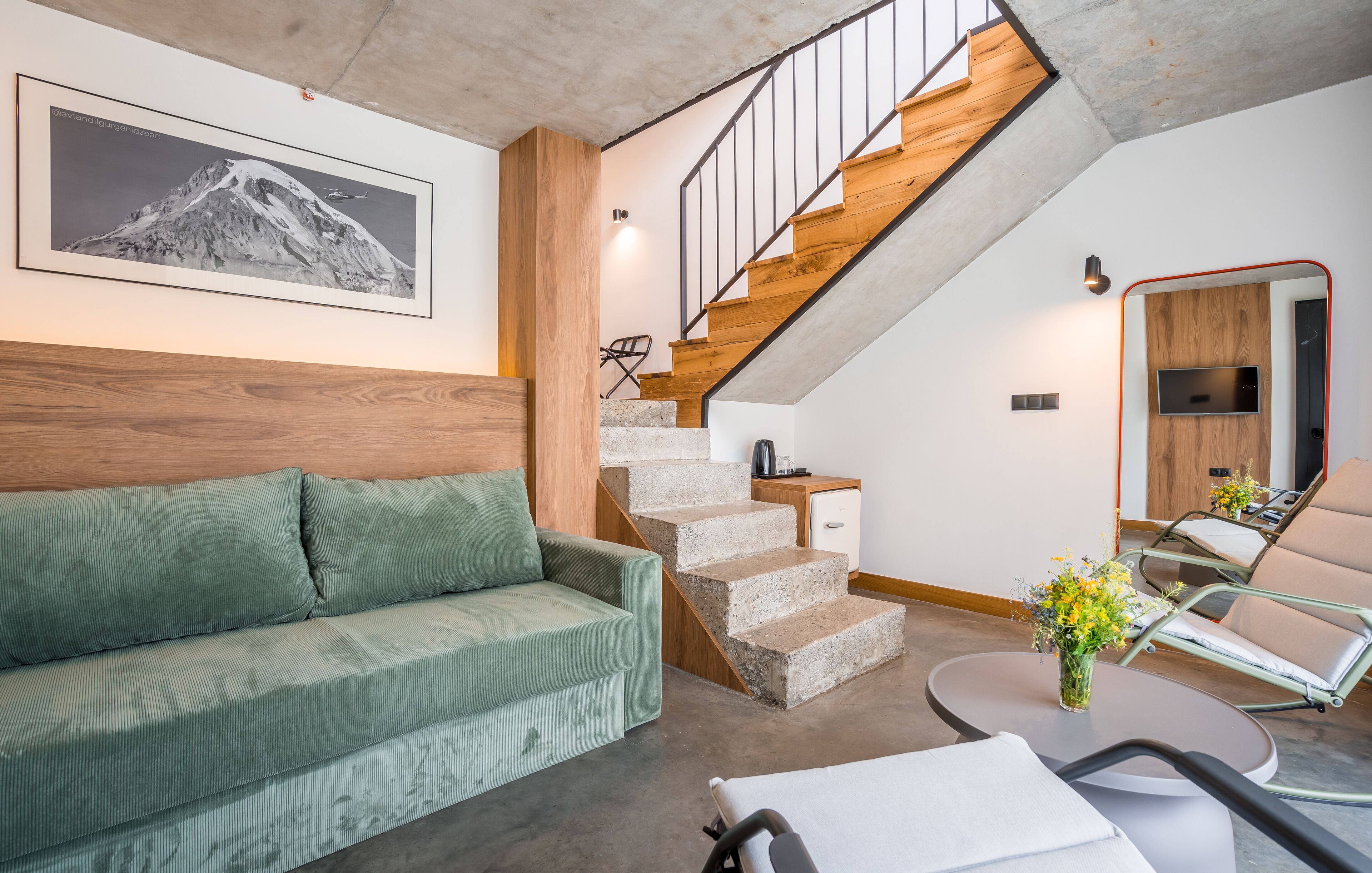Uma sala de estar moderna com um sofá verde, uma mesa de centro redonda, uma cama de hospital e uma escada com acabamento em madeira.