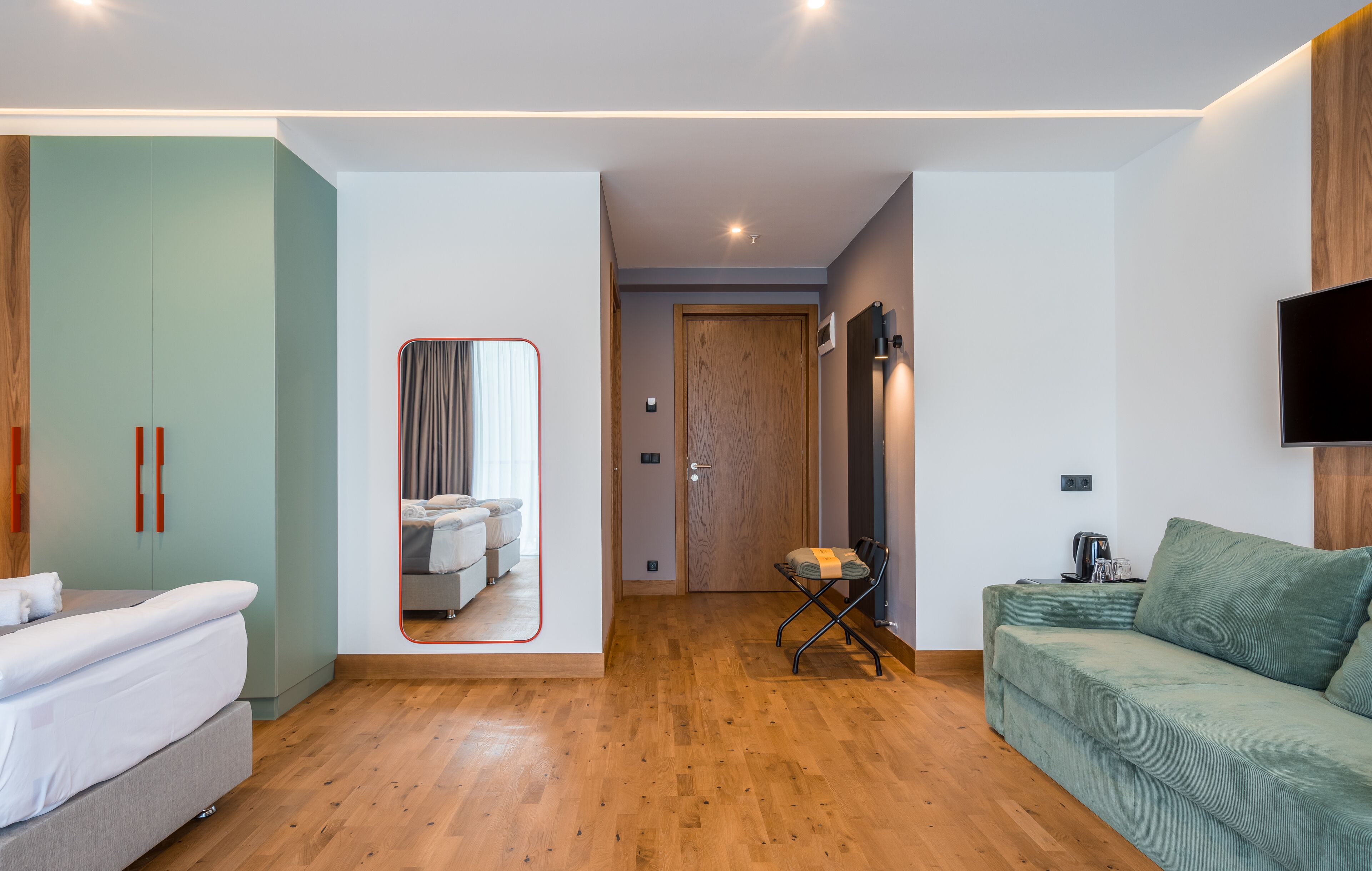 Ein modernes Hotelzimmer mit Holzboden, einem grünen Sofa, einem Bett und einem an der Wand befestigten Fernseher.