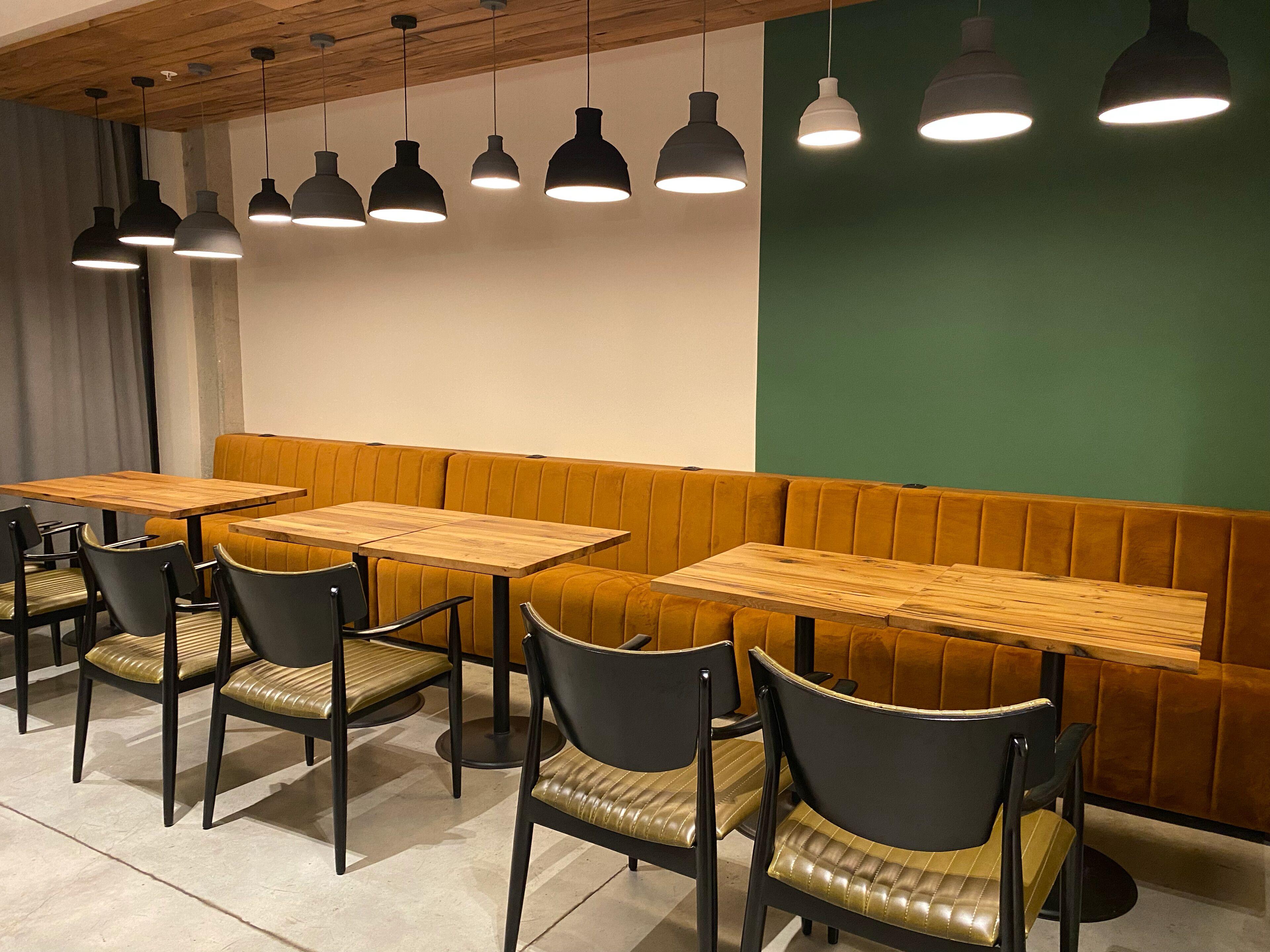 A restaurant interior with wooden tables and black chairs, a green accent wall, and pendant lighting.
