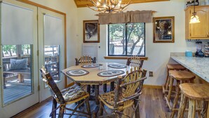 Un interior rústico con una mesa de comedor de madera rodeada de sillas variadas, una zona de cocina con un taburete de bar y una araña.