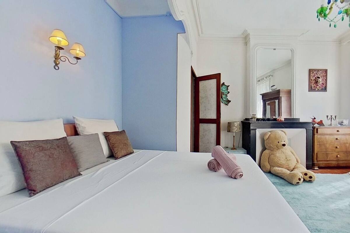 A neatly made bed with pillows, a teddy bear, and a lamp on the wall.