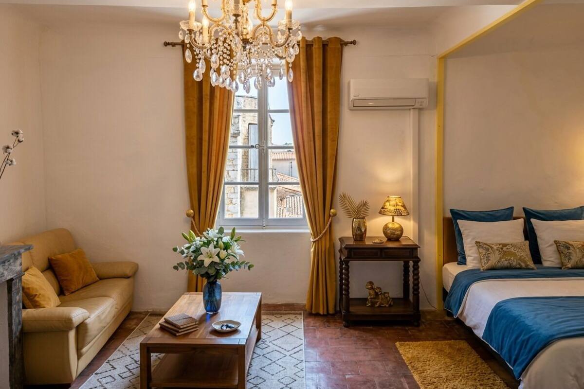 A room with a bed, a sofa, a chandelier, a window with curtains, a small table with a lamp, and an air conditioning unit.