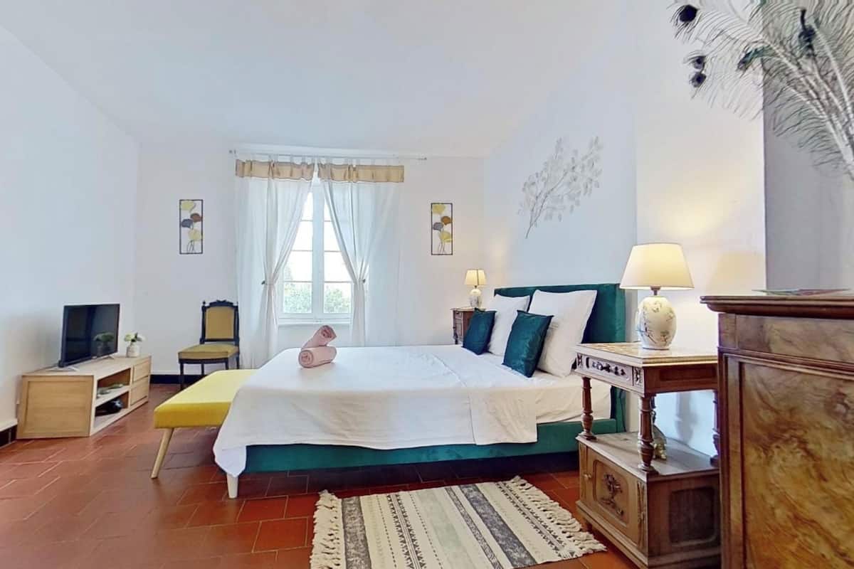 A bedroom with a bed, bedside tables, a TV, a window with curtains, and a wall decoration.