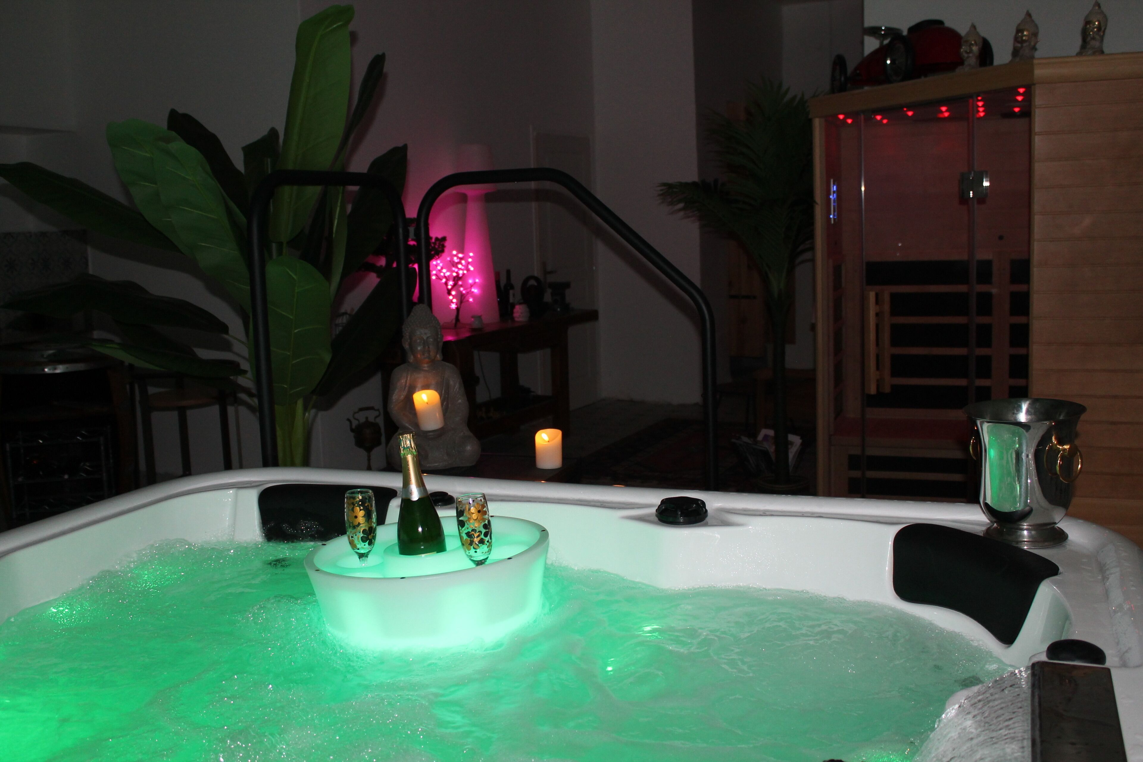 Un bain à remous agrémenté d’une bouteille décorative et d’une bougie, entouré de plantes et d’un meuble en bois.